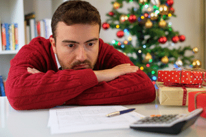 A man in a red jumper looking at his desk. There is a calculator, a pen and piece of paper on the desk. There is a Christmas tree in the background
